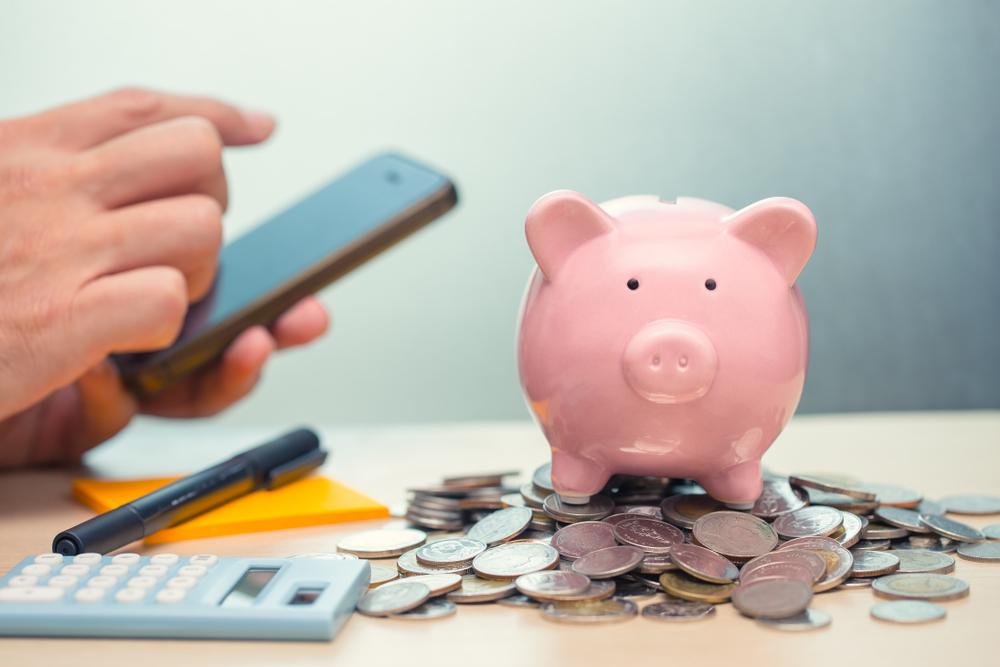 hand holding a mobile phone next to a piggy bank and cash